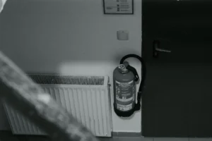Czarno-białe zdjęcie gaśnicy zamontowanej na ścianie obok kaloryfera i drzwi na klatce schodowej starego budynku.
